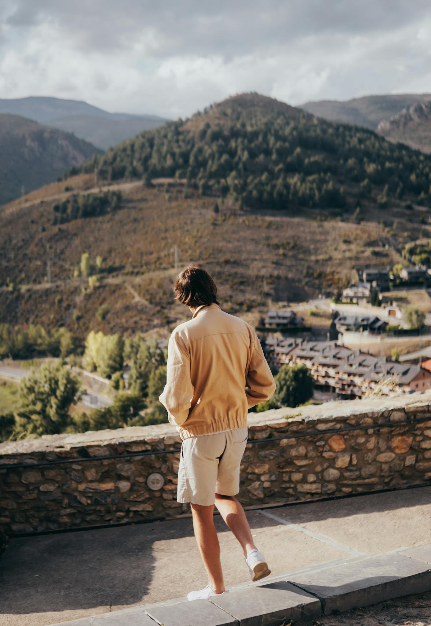 Man walking along a stone wall with a scenic mountain view on a bright day.